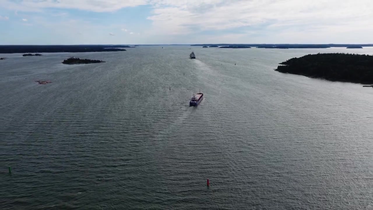 Lille fragtskib navigerer gennem finsk skærgård. Fartøjet passerer en smal sejlrende tæt på kystlinjen. Stigende luftfoto agterfra afslører passagen forude