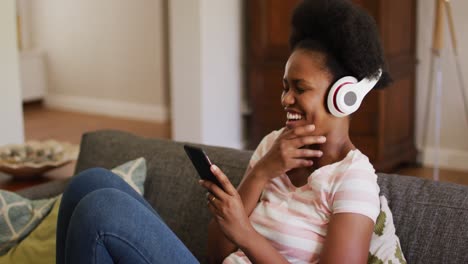 Feliz-Mujer-Afroamericana-Usando-Auriculares-Relajándose-En-El-Sofá-Usando-Un-Teléfono-Inteligente-Riendo