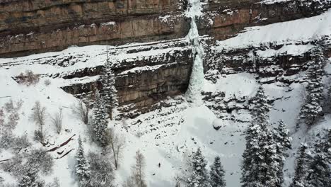 aerial view of lonely person walking on snow under cliffs with frozen waterfall on cold winter day, tilt up drone shot