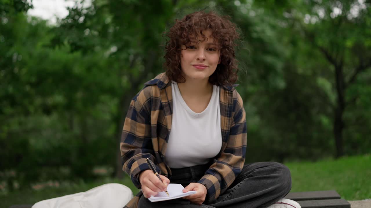 Portrait of a brunette student girl with curly hair in a brown shirt sitting on a bench and making notes in a notebook in the park