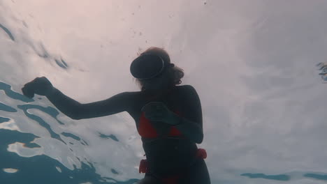 Cinematic-bottom-up-shot-of-a-beautiful-woman-in-an-orange-bikini-and-vintage-diving-goggles-swimming-close-to-the-water-surface-in-4K,-120-FPS,-Slomo