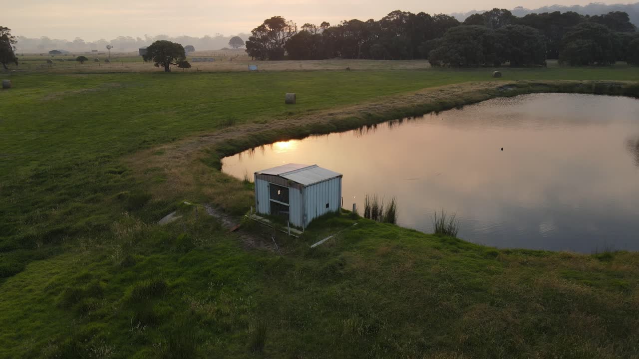 Droneoptagelse i kredsløb over et lille hus og en sø under en tåget solnedgang på en gård.