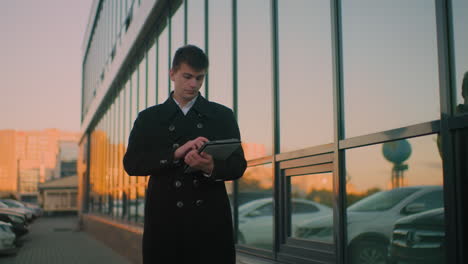 business mogul in black coat walking near glass building, focusing on tablet, reflections on glass show blurred building and parked cars, emphasizing modern city life