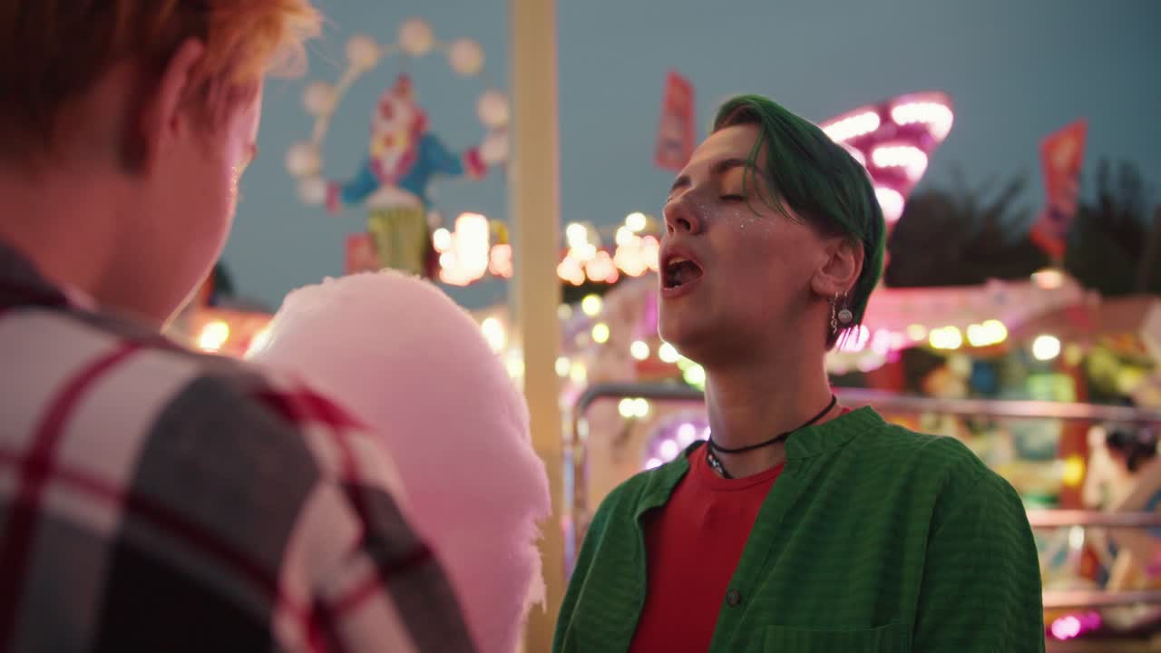 A blonde girl with a short haircut in a plaid shirt feeds her girlfriend with a short green haircut in a Green shirt and also passes a piece of pink cotton candy with her mouth during their date in a bright and glowing amusement park