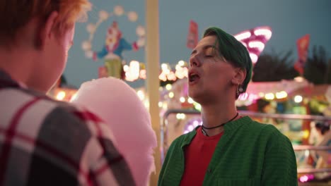 a blonde girl with a short haircut in a plaid shirt feeds her girlfriend with a short green haircut in a green shirt and also passes a piece of pink cotton candy with her mouth during their date in a bright and glowing amusement park