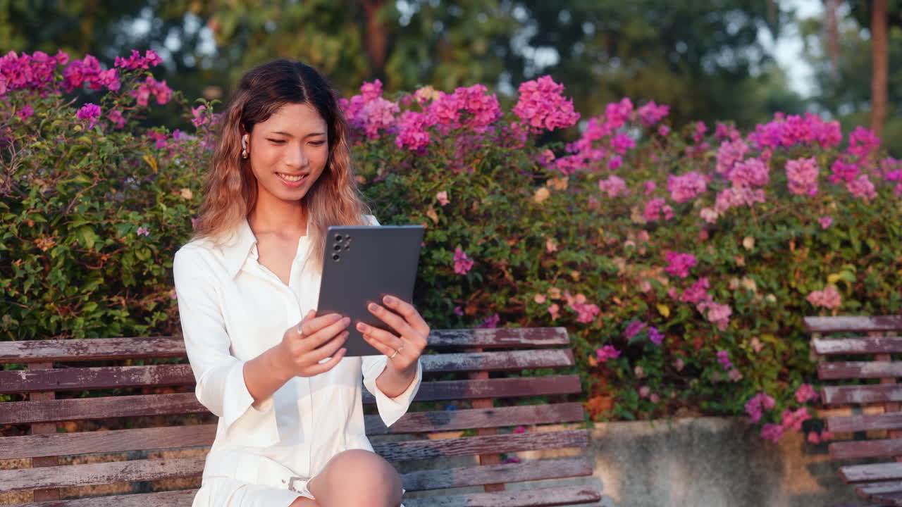 Young Asian Woman Using Tablet on a Park Bench with Pink Flowers