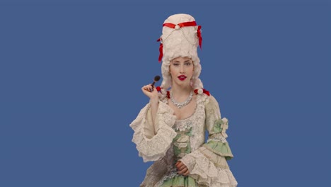 portrait of courtier lady in vintage dress and wig is applying powder to her face using a makeup brush. young woman posing in studio with blue screen background. close up. slow motion ready 59.94fps