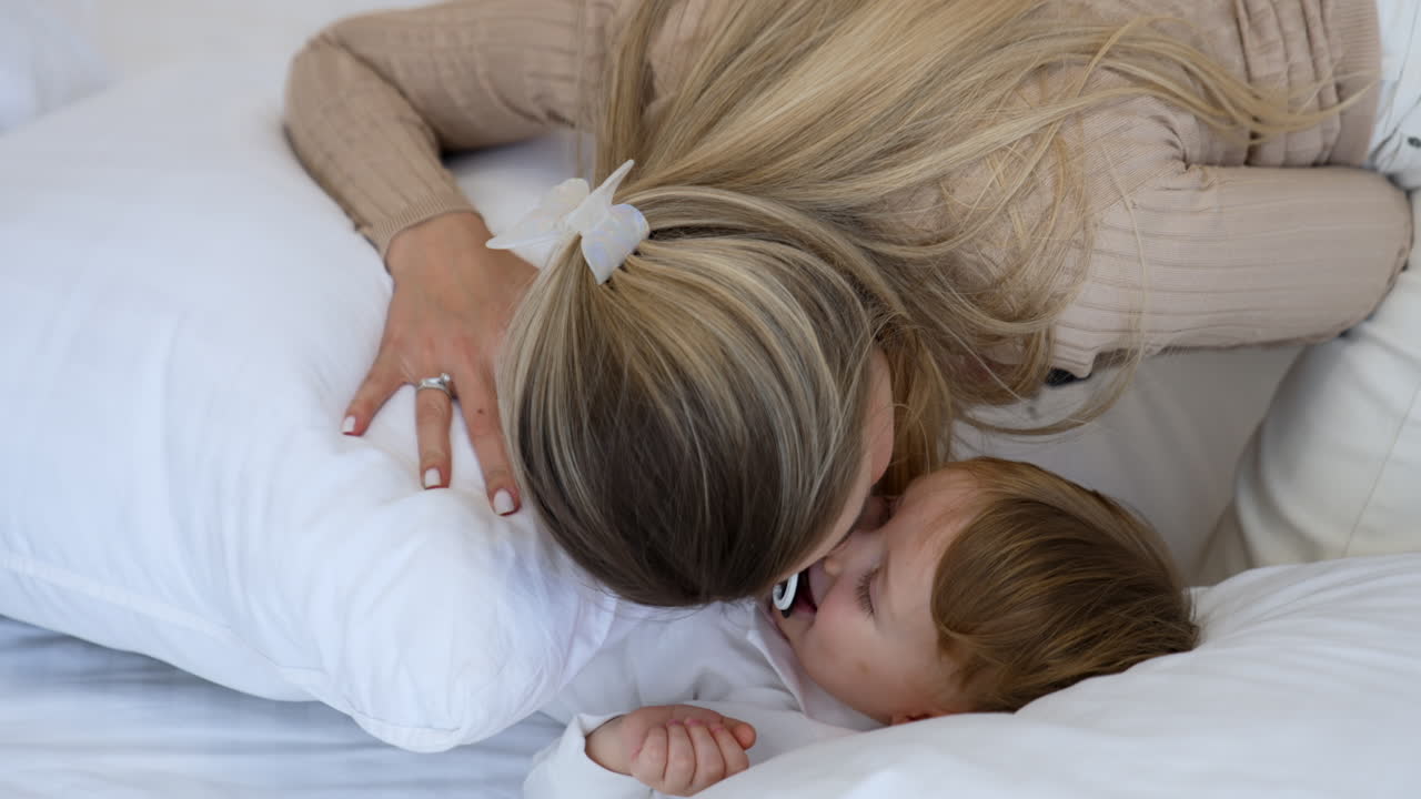 Mom puts a pillow on her little baby boy sucking a pacifier. Loving mother kisses her adorable child on the cheek.