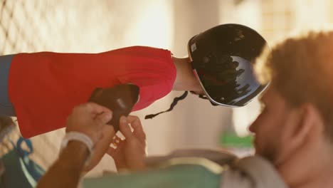 Vertical-video-of-a-dad-with-curly-hair-in-a-Green-T-shirt-helping-his-little-blond-son-with-blue-eyes-in-a-red-T-shirt-put-on-protective-clothing-before-riding-a-skateboard-in-the-park