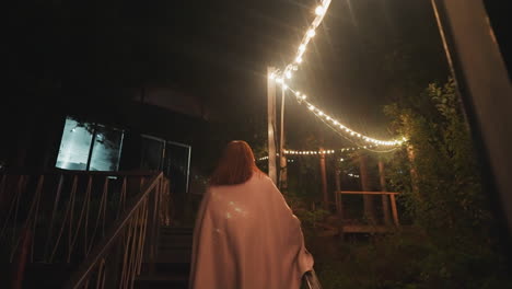 young woman in blanket climbs stairs. lonely woman returns home from evening walk to wooden house. staircase illuminated with decorative bulbs