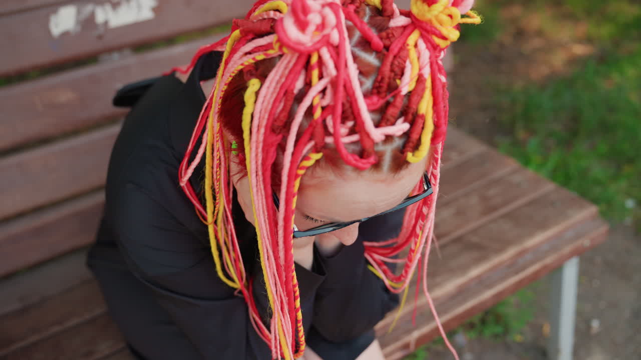 분홍색과 주황색 땋은 머리를 한 백인 구직자가 나무 벤치에 움츠린 채 앉아 머리를 감싸 쥐고 불안한 표정을 짓고 있어요. 클로즈업 탑다운 앵글로 긴장감을 포착했으며, 주변에는 공원의 나무들과 여름 햇살이 비치고 있어요.