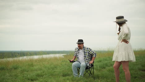 a woman in a white dress stands with her hands on her hips, watching as an artist in a checked shirt sits down on a chair in a grassy field