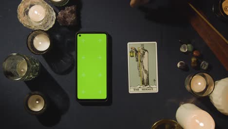 Draufsicht-Einer-Person,-Die-Neben-Der-Einsiedlerkarte-Auf-Dem-Tisch-Tarotkarten-Liest,-Mit-Einem-Mobiltelefon-Mit-Grünem-Bildschirm