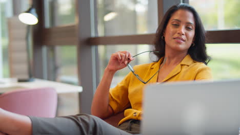 Mature-Businesswoman-With-Glasses-Relaxing-With-Feet-On-Desk-In-Office-Using-Laptop