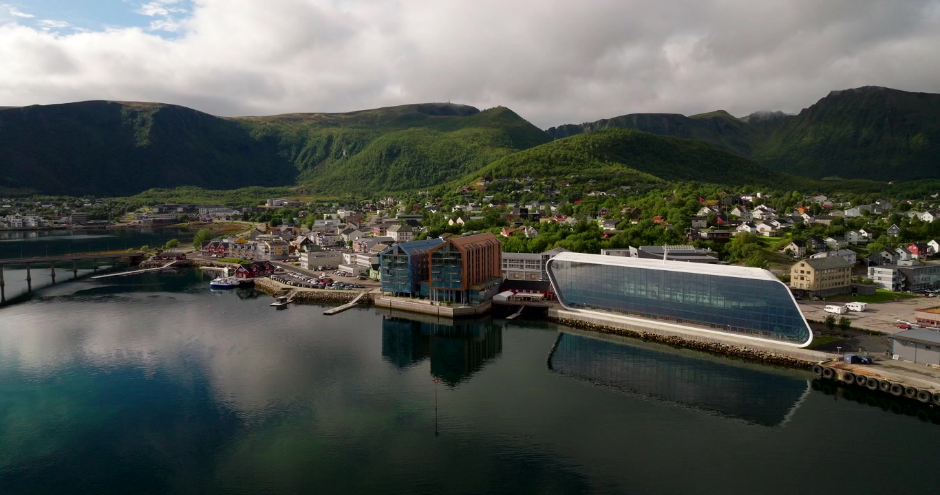 Stokmarknes, Norge, med Hurtigrutemuseets strandnära komplex, hamn och omgivande berg speglade i lugna vatten, filmisk flygbild.
