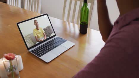 sección media de un hombre afroamericano bebiendo vino mientras hace una videollamada en una computadora portátil en casa