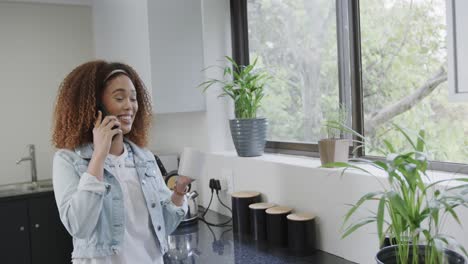 Happy-biracial-woman-talking-on-smartphone-and-drinking-coffee-in-kitchen,-slow-motion