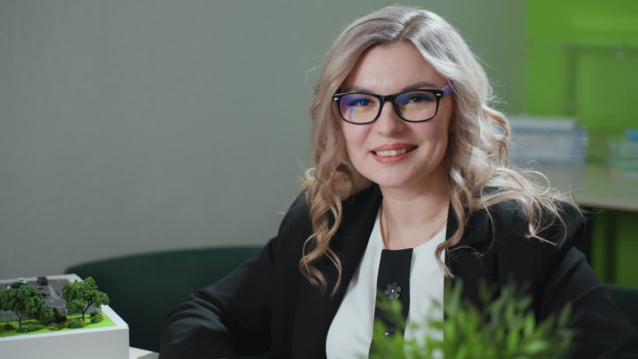 realtor with curly blonde hair and glasses looks up from laptop with elegant smile in modern office, surrounded by architectural model, desktop supplies, greenery, and professional workspace