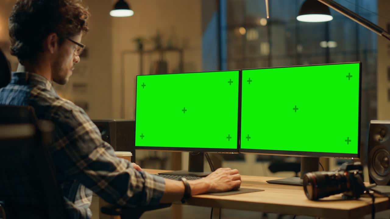 por encima del hombro: diseñador creativo sentado en su escritorio utiliza una computadora de escritorio con dos pantallas de maquetación verdes. empleado de oficina profesional trabajando tarde en la noche en su estudio