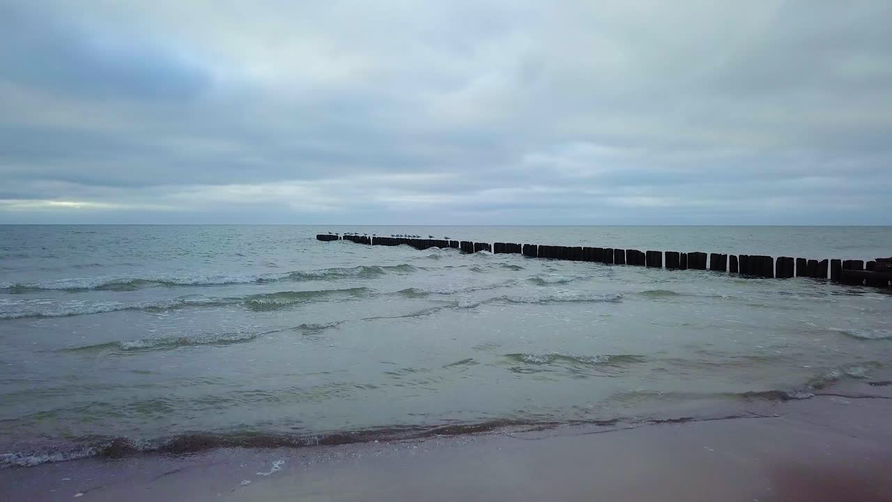 흐린 날 발트해 연안의 공중 전망, 오래된 목조 부두, 하얀 모래 해변, 해안에 부서지는 낮은 파도, 기후 변화, 낮은 광각 드론 샷