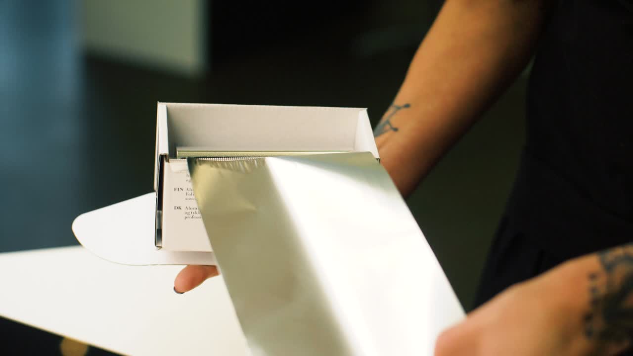 Tattooed hairdresser is cutting sheets of foil from dispenser. Getting ready to dye clients hair. Slow motion.