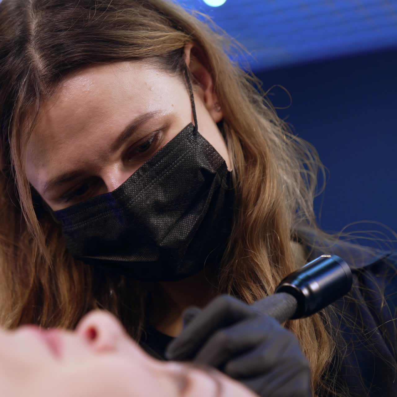 Long haired Caucasian beautician wearing black mask. Cosmetologist does permanent make up of eyebrows. Close up