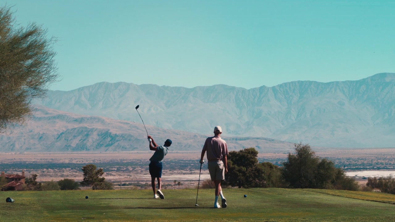 Two golfers tee off with a driver above a desert valley and mountain backdrop in slow motion, suited to golf getaway marketing and friends-playing lifestyle stories.