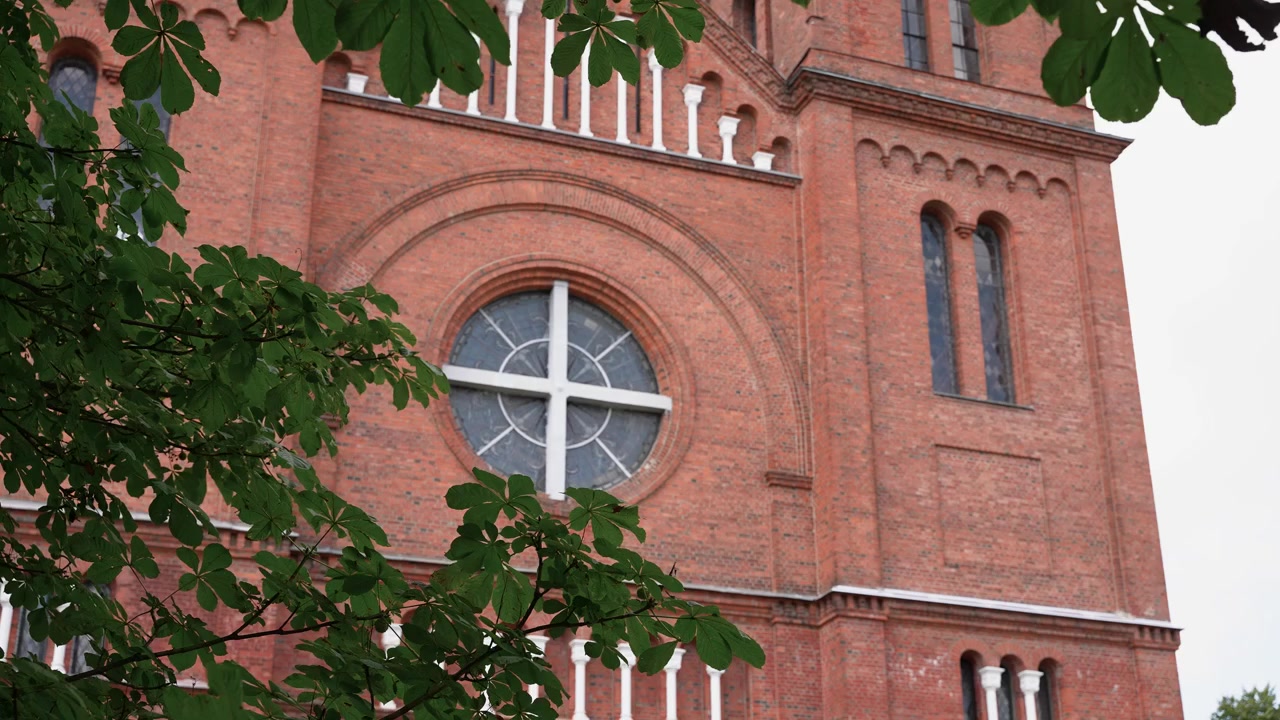 Imagem detalhada da fachada de tijolos vermelhos da Igreja de São João Batista em Plungė, Lituânia, com vitral redondo e folhas de árvores verdes exuberantes sob luz diurna suave. Sem pessoas