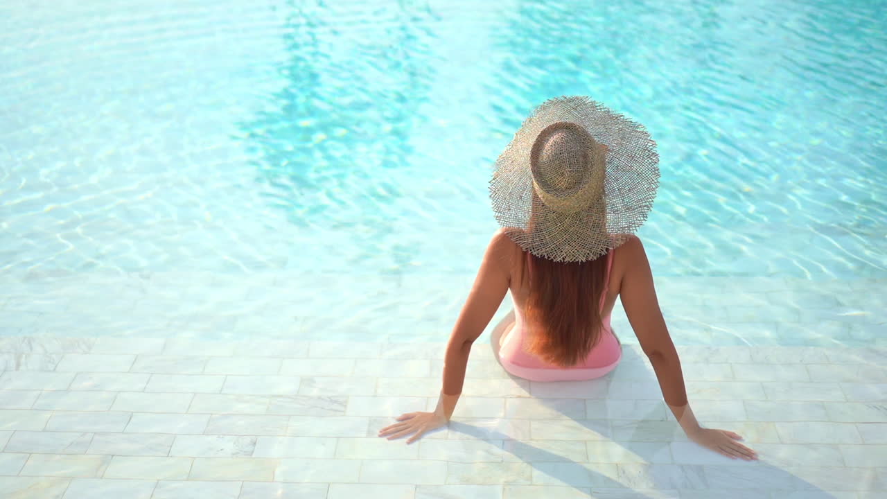 Back of Sexy Female in Swimsuit and Summer Hat Sitting on Pool Edge on Hot Day. Fashion and Luxury Travel Concept, Static Full Frame