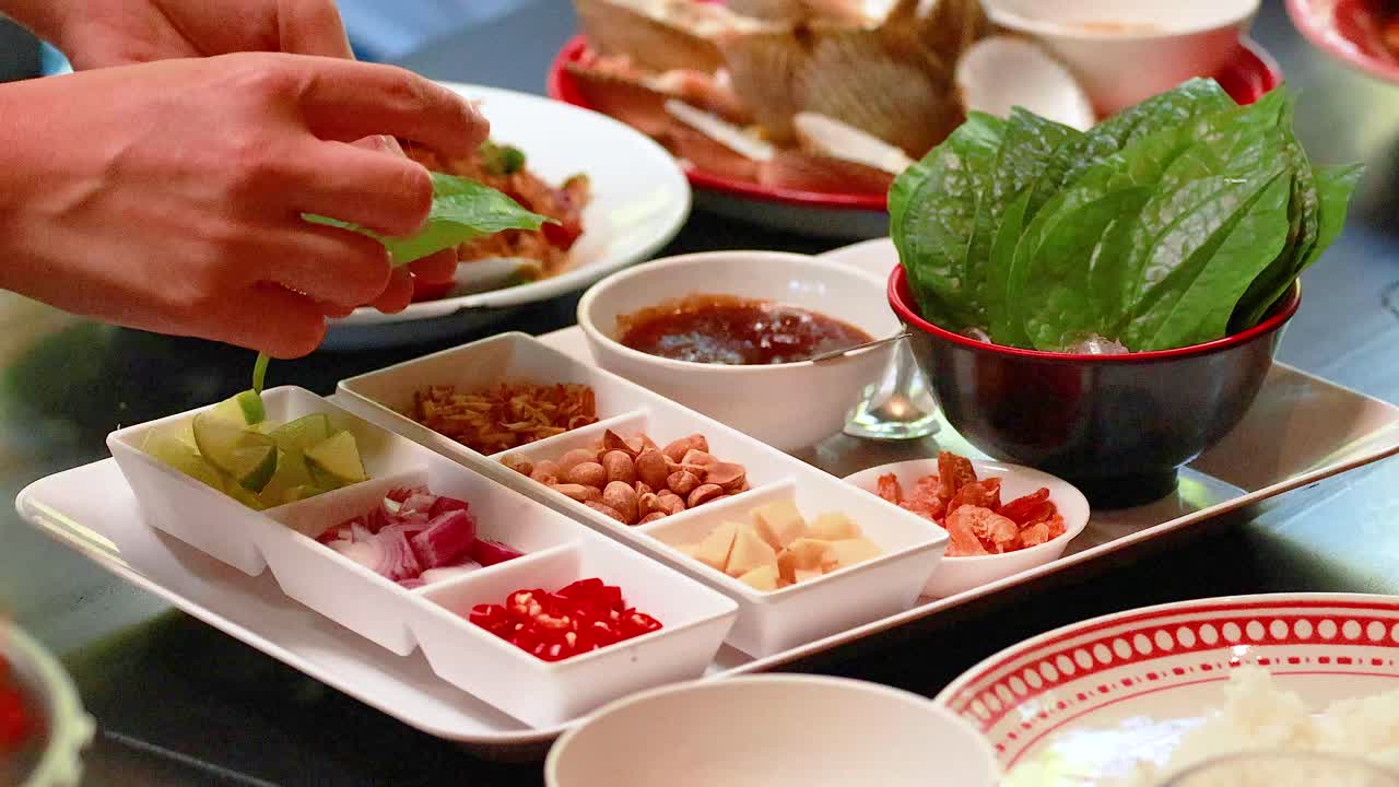 Hands assembling Miang Kham with fresh ingredients on a table in Phuket. Bright lighting enhances the vibrant colors of the dish
