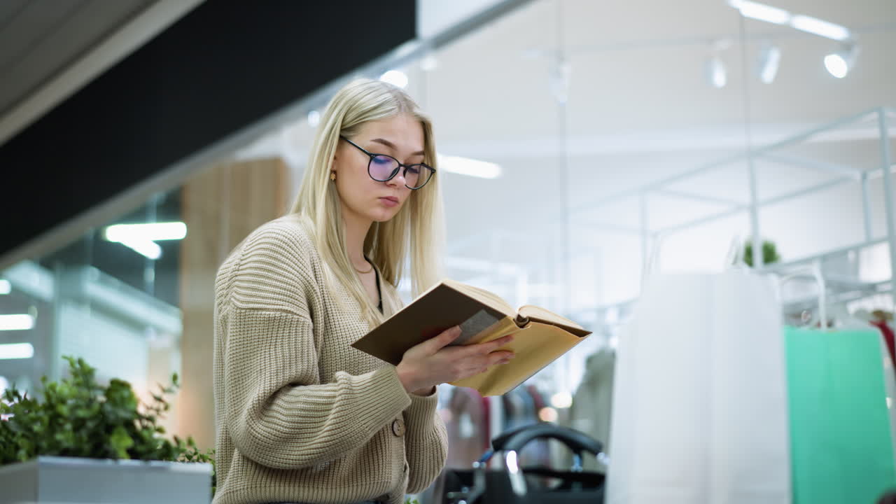 joven con gafas se centra en el libro, volviendo las páginas pensativamente con flor decorativa verde, ropa visible a través del vidrio de la tienda, bolsa de compras blanca y menta, bolso negro cerca