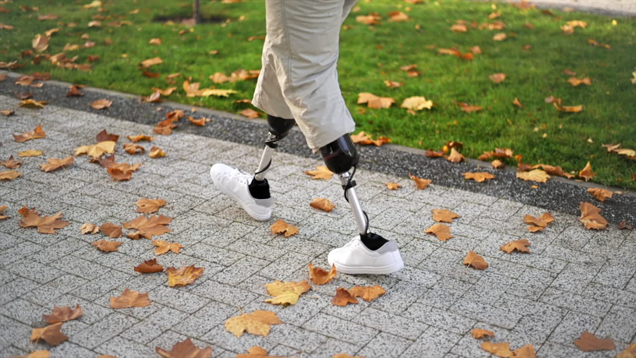 Slow motion view of a walking man with prosthetic legs and white sneakers in a park. Fallen yellow leaves on the ground