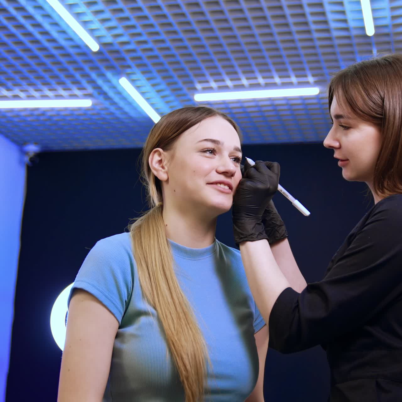 Long-haired brunette cosmetologist draws lines near the eyebrows of a client. Preparation for eyebrow permanent make-up. Low angle view