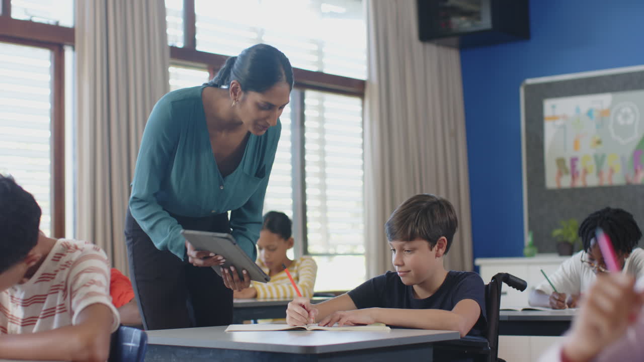 Assisting student with tablet in classroom, teacher engaging in school discussion