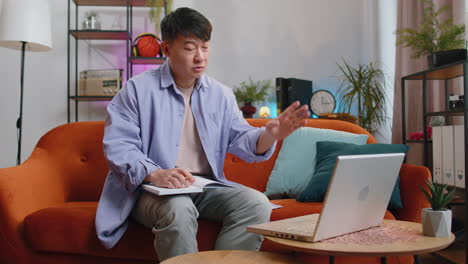 asian handsome man at home making conference video call talking to webcam, making notes on notebook