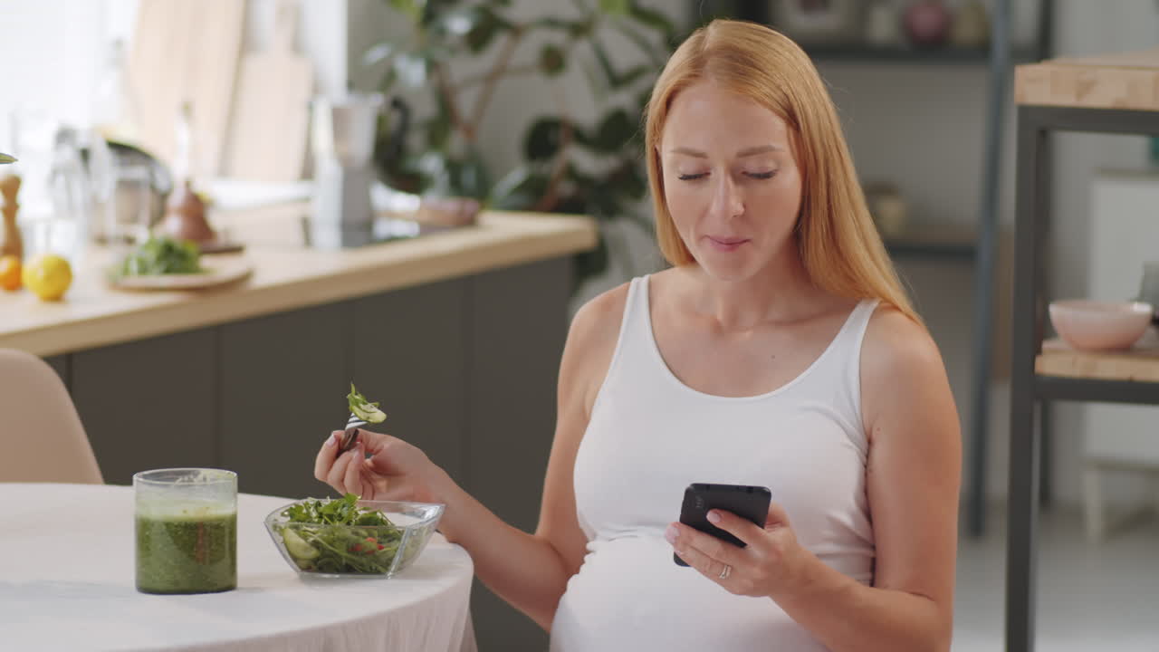 Pregnant Woman Eating Healthy Salad and Using Phone in Kitchen