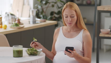 pregnant woman eating healthy salad and using phone in kitchen