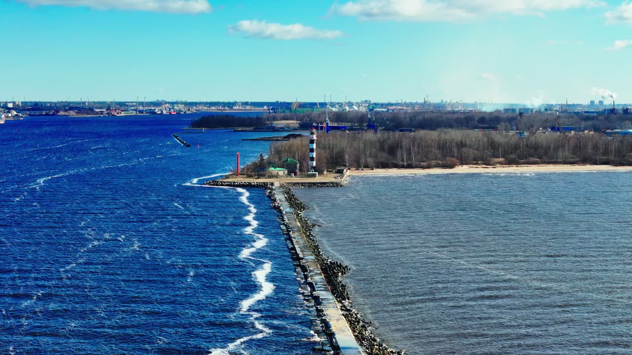 A long stone jetty stretches into vivid blue Baltic Sea, ending at a tall black and white lighthouse dividing choppy waves from muted estuary waters and sandy coastline.