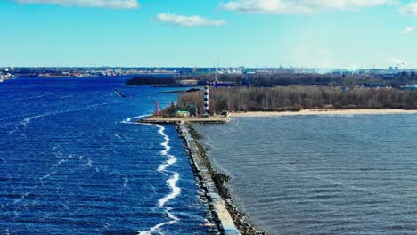 a long stone jetty stretches into vivid blue baltic sea, ending at a tall black and white lighthouse dividing choppy waves from muted estuary waters and sandy coastline.