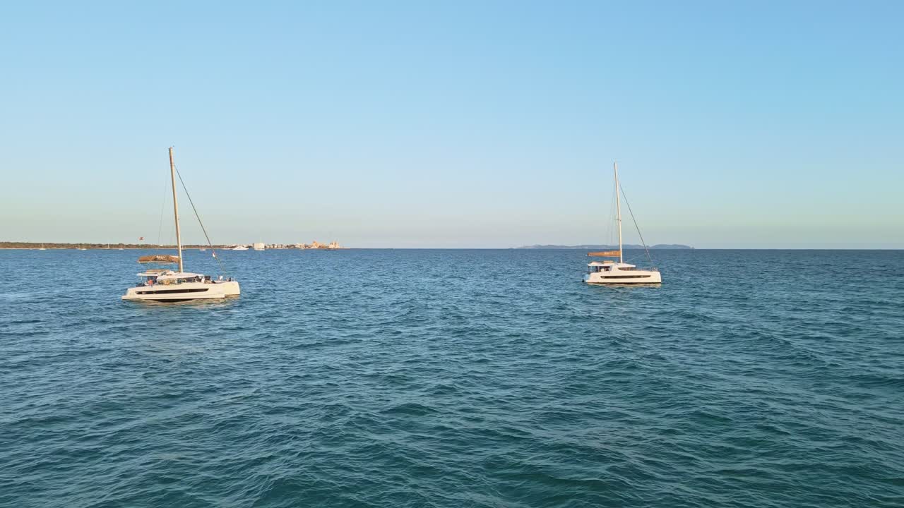 A dynamic fly-through reveals two identical catamarans anchored off Es Trenc, Mallorca, as they float on the clear blue waters of the Mediterranean.