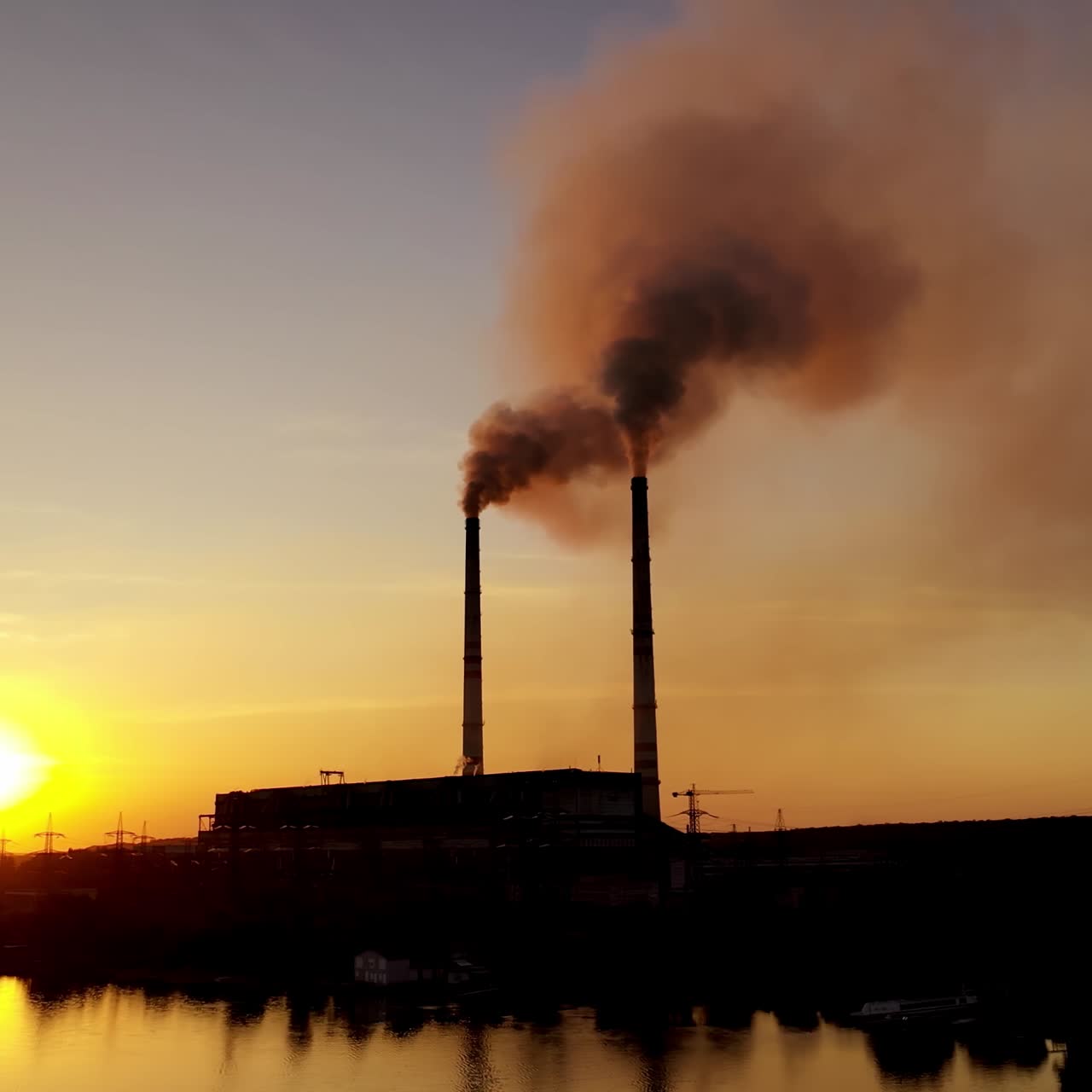 Dark power plant at the bank of the evening river. Industrial pipes of manufacturing with dark smoke at sunset. Aerial view.