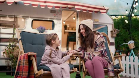 Happy-little-blonde-girl-in-a-pink-hoodie-communicates-with-her-mom,-a-brunette-woman-in-a-plaid-shirt-and-pink-pants,-during-her-vacation-in-a-camp-near-a-trailer-outside-the-city-in-summer