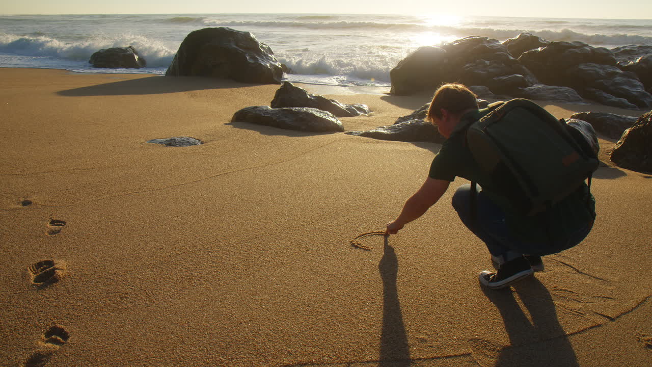 Young male drawing a heart in the sand of the beach at sunset