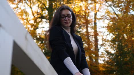 Young-Woman-in-Casual-Outfit-Sitting-on-Fence-at-Public-Park-on-Autumn-Day,-Slow-Motion-of-Teenage-Girl-Touching-Hair-With-Fall-Colors-in-Background,-Focus-In