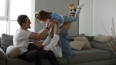 happy family mom and dad having fun with little girl on cozy home grey sofa. little daughter imitates flying being on her