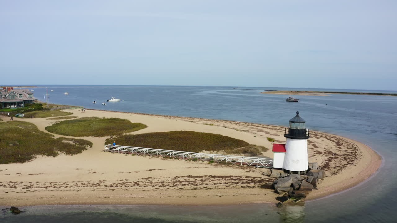 Drone che orbita a sinistra attorno al Faro di Brant Point a Nantucket, Massachusetts, mostrando la penisola sabbiosa, le imbarcazioni di passaggio e le calme acque del porto in un'ampia ripresa aerea costiera.