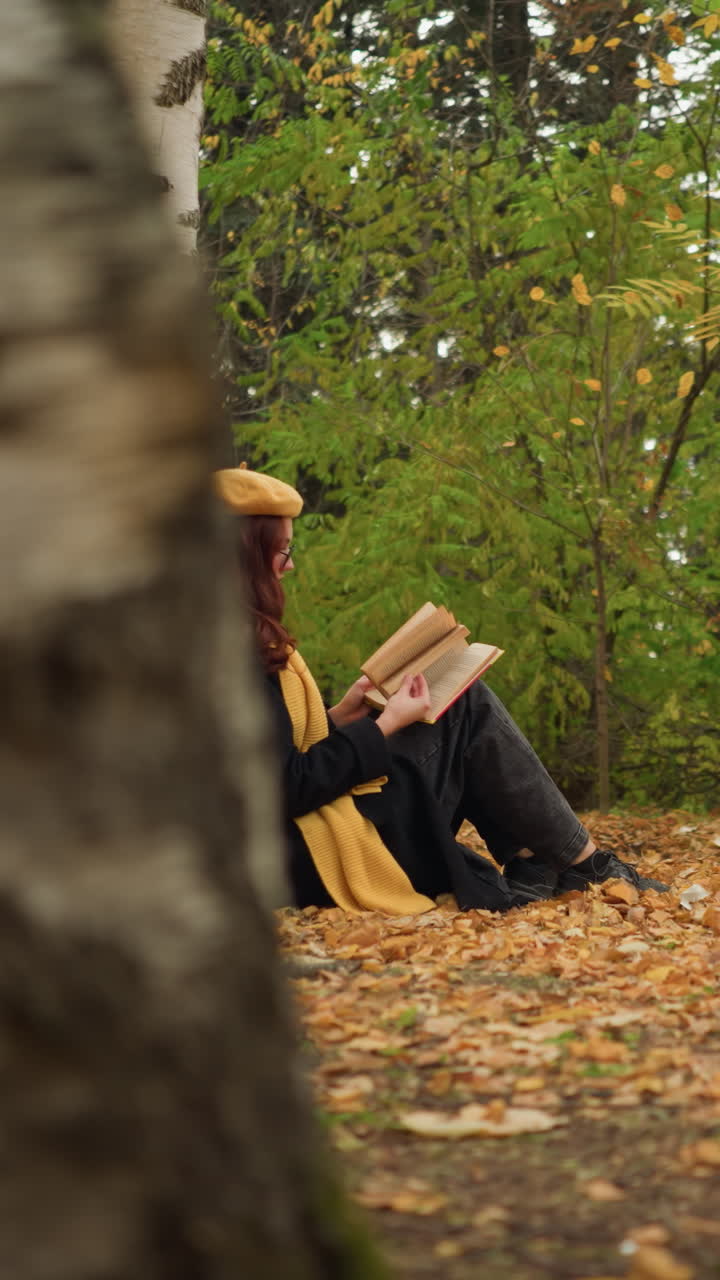 joven con boina amarilla y cabello rojo cayendo por el hombro leyendo cuidadosamente un libro en la mano mientras estaba sentada contra un abedul, rodeada de hojas de otoño en paz y soledad