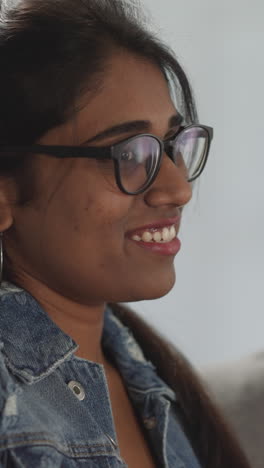 indian woman with ponytail smiles watching comedy movie with friend. young lady in glasses sits on sofa enjoying tv after work at home closeup