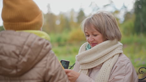 little child playfully reaches out holding yellow leaf to touch mum head as mum smiles warmly and moves backward on autumn park bench surrounded by blurred foliage and seasonal ambience