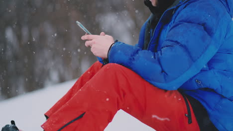 male snowboarder sitting on the snow taking photos on the phone beautiful scenery of the resort for social networks